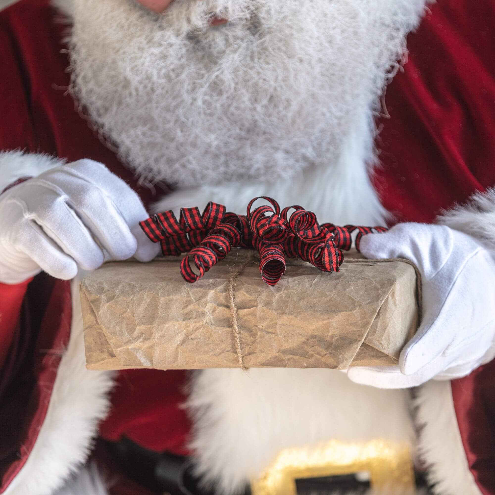 A man in a Santa suit unwrapping a Christmas present wrapped in brown paper and a red ribbon.