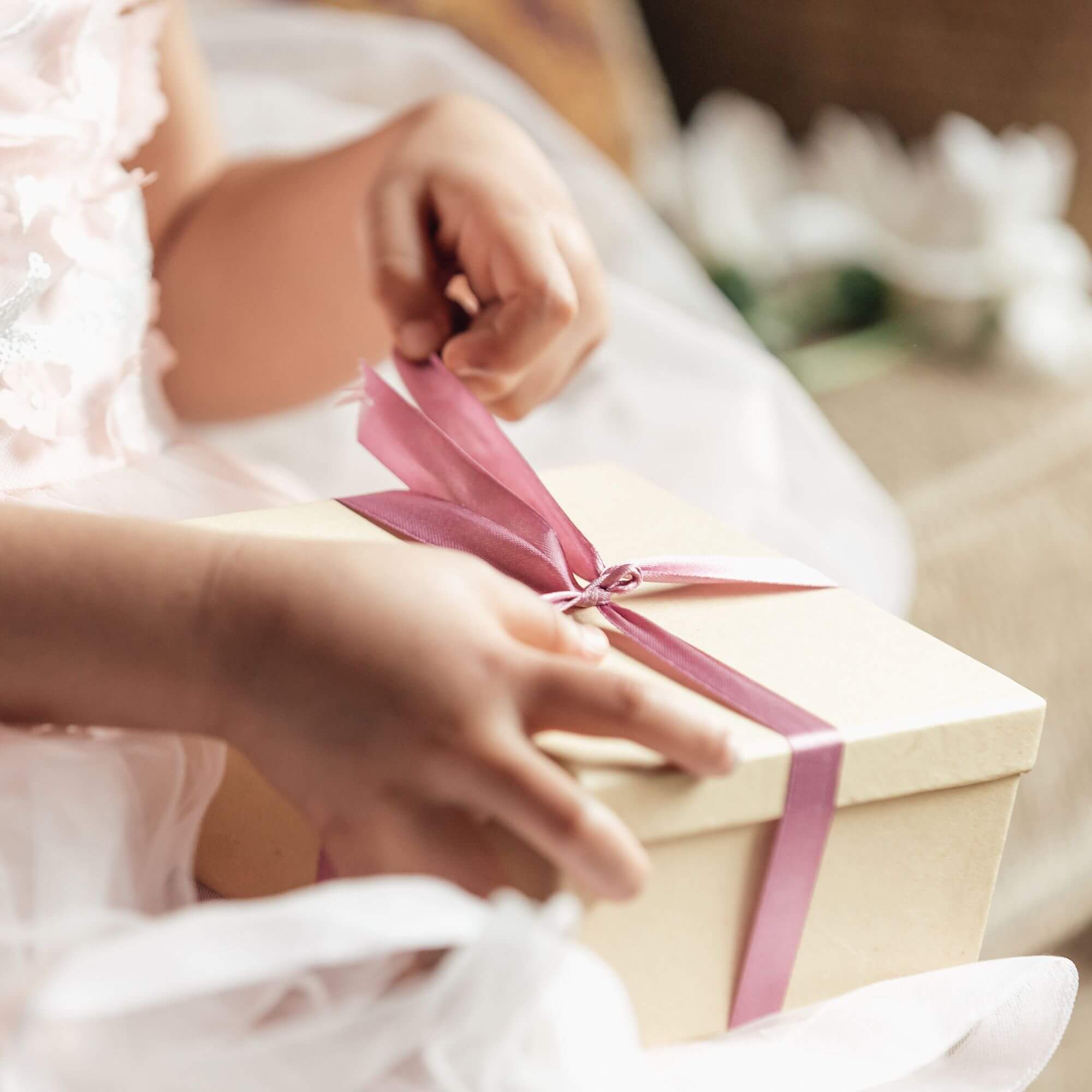 A child in a white dress opens a Christmas gift in a brown box and a pink ribbon.